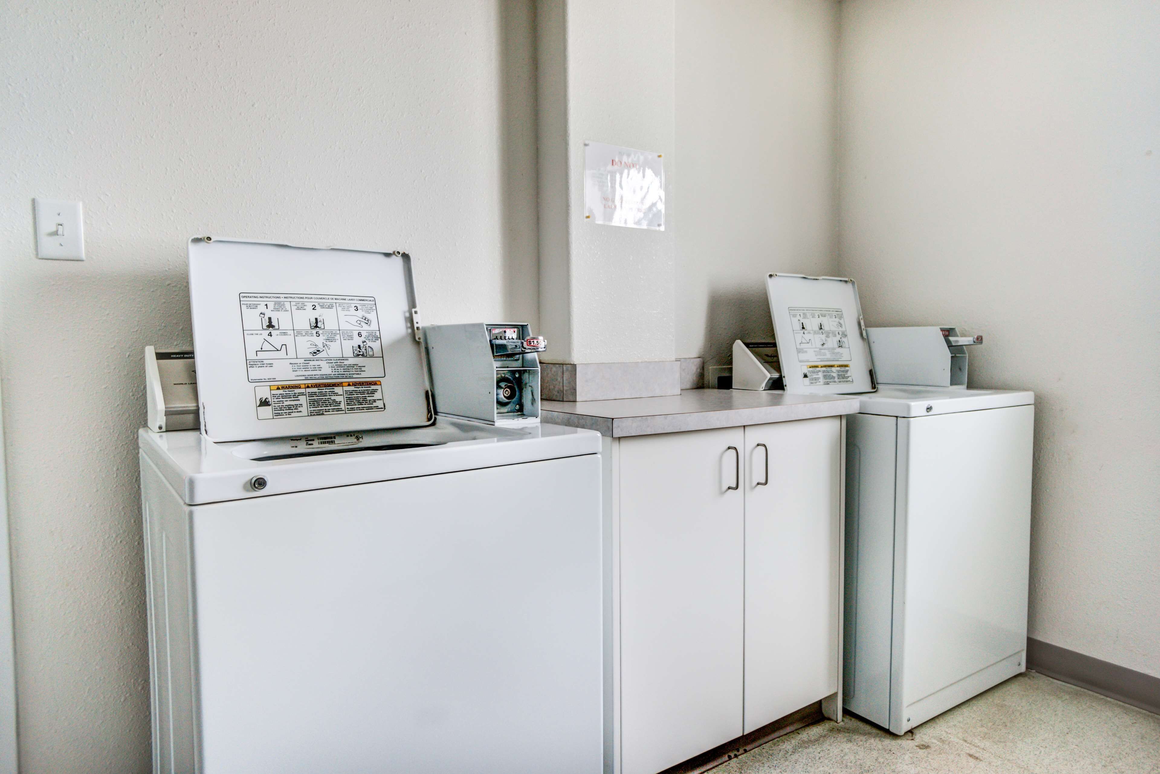 Een wasserette met twee witte wasmachines en drogers, een bedieningspaneel en een bericht op de muur.