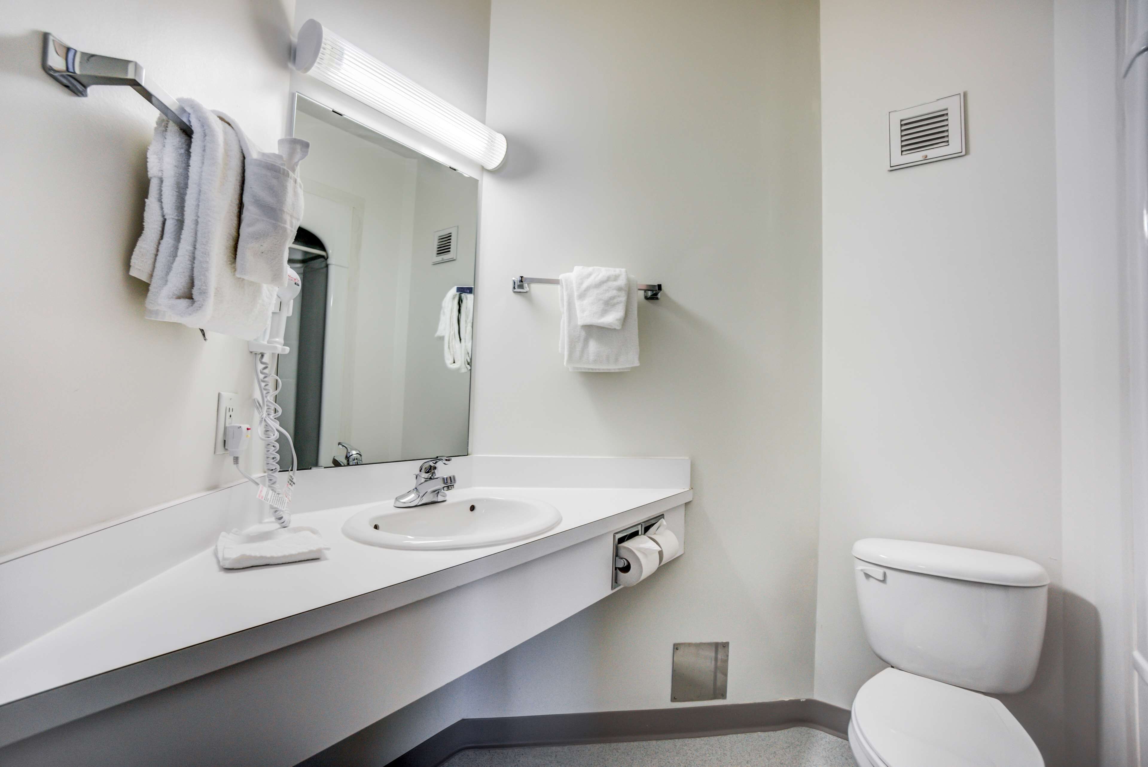 Une salle de bain moderne avec un lavabo blanc, un miroir et des toilettes. Des serviettes sont suspendues sur un porte-serviettes et un distributeur de serviettes est également présent.
