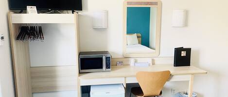A compact hotel room with a wooden desk, a microwave, a small refrigerator, a chair, a TV mounted on the wall, and a mirror.