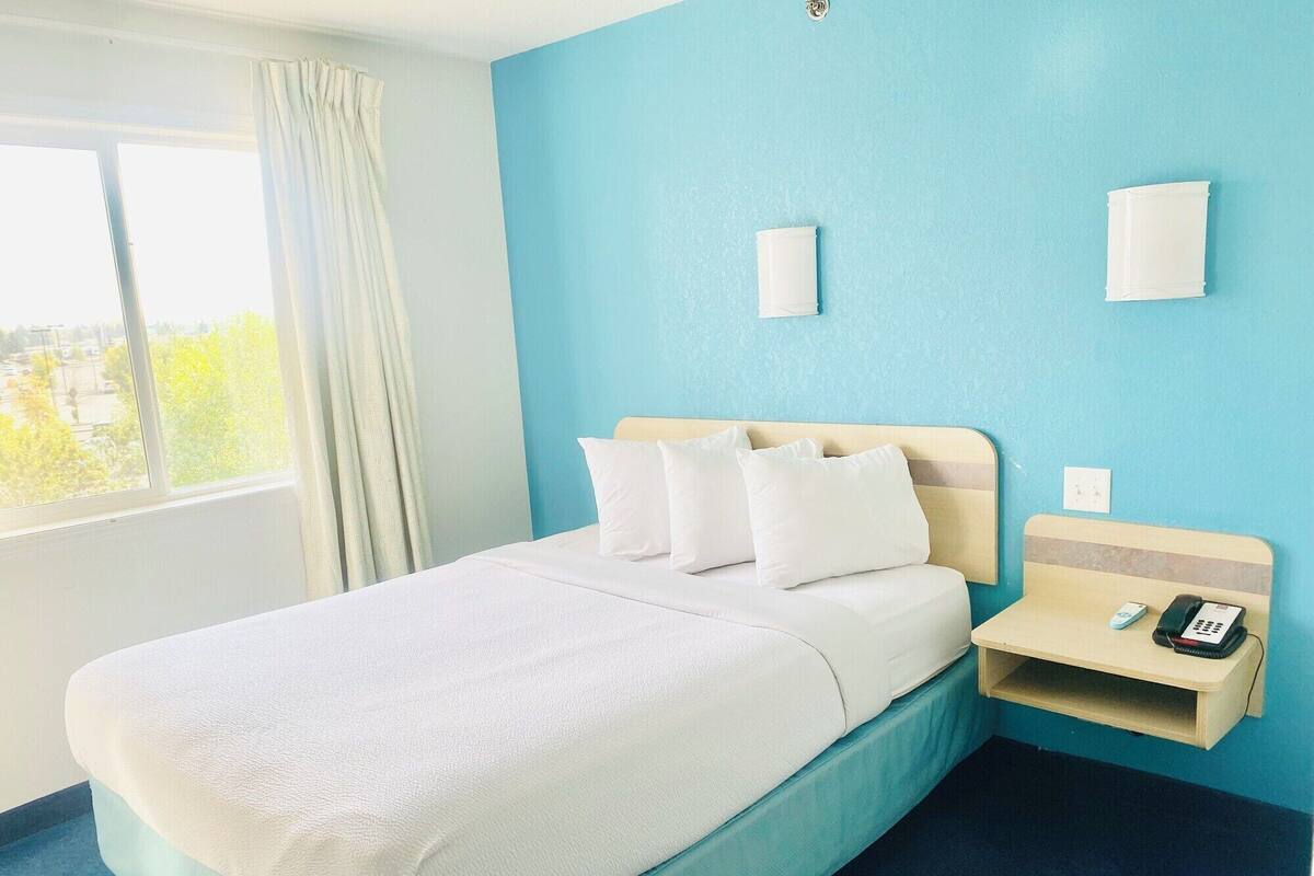 A hotel room with a bed, a bedside table with a phone, and a window with curtains.