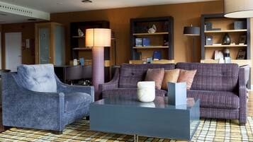 Una sala moderna con un sillón azul, un sofá morado y una mesa de centro de vidrio. Hay dos lámparas de pie, una estantería y una puerta al fondo.