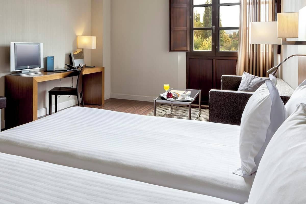 Une chambre d’hôtel avec un lit, un bureau avec un ordinateur, une chaise, une petite table avec une boisson et une fenêtre avec des rideaux.