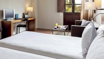 Une chambre d’hôtel avec un lit, un bureau avec un ordinateur, une chaise, une petite table avec une boisson et une fenêtre avec des rideaux.
