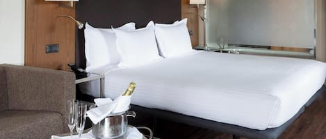 Habitación de hotel con una cama grande, una mesita de noche con una botella de champán, copas y una bandeja, un cabecero de madera y una zona de ducha.