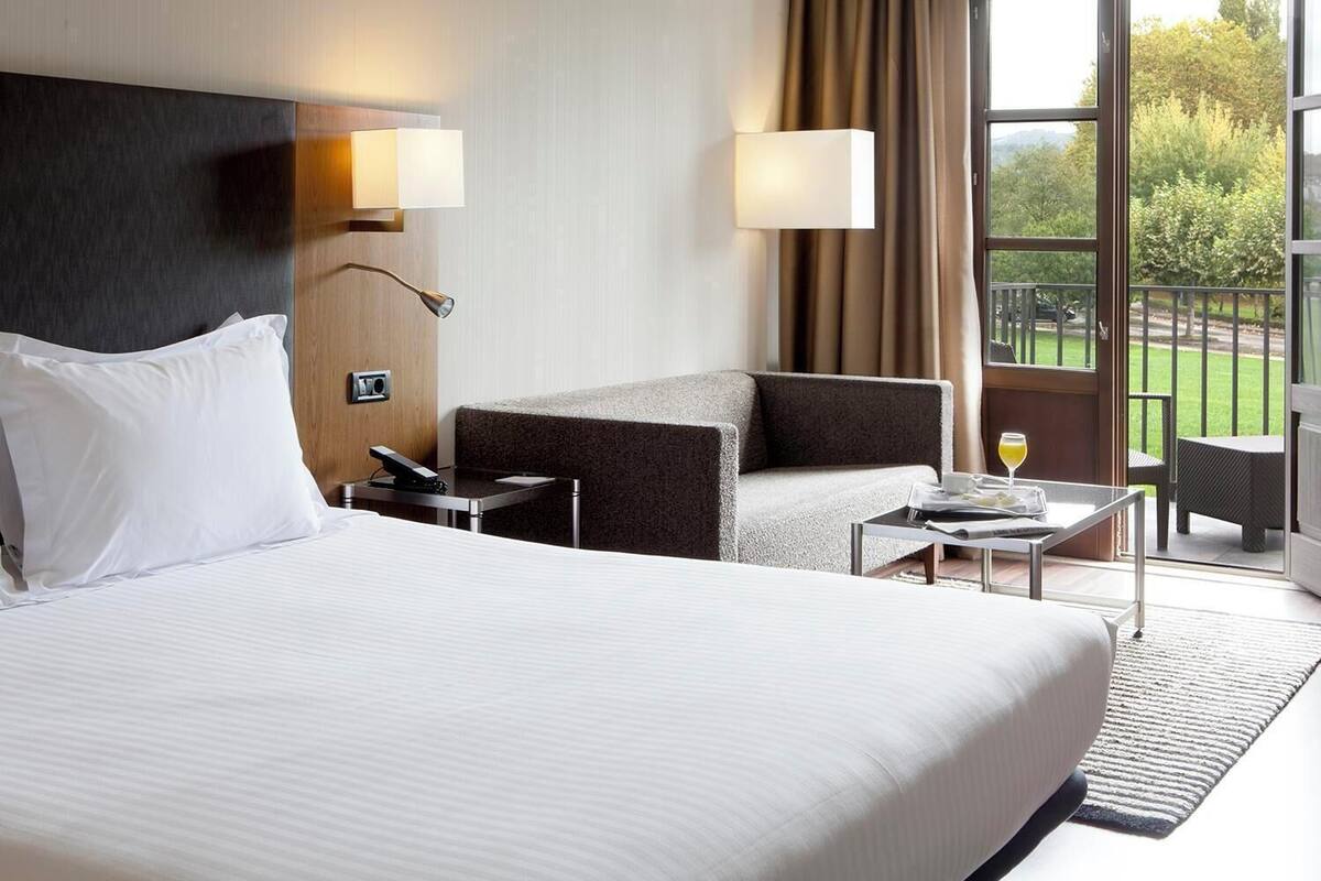 Une chambre d’hôtel avec un grand lit, un bureau, une chaise, une petite table avec une boisson, et une vue sur l’extérieur.