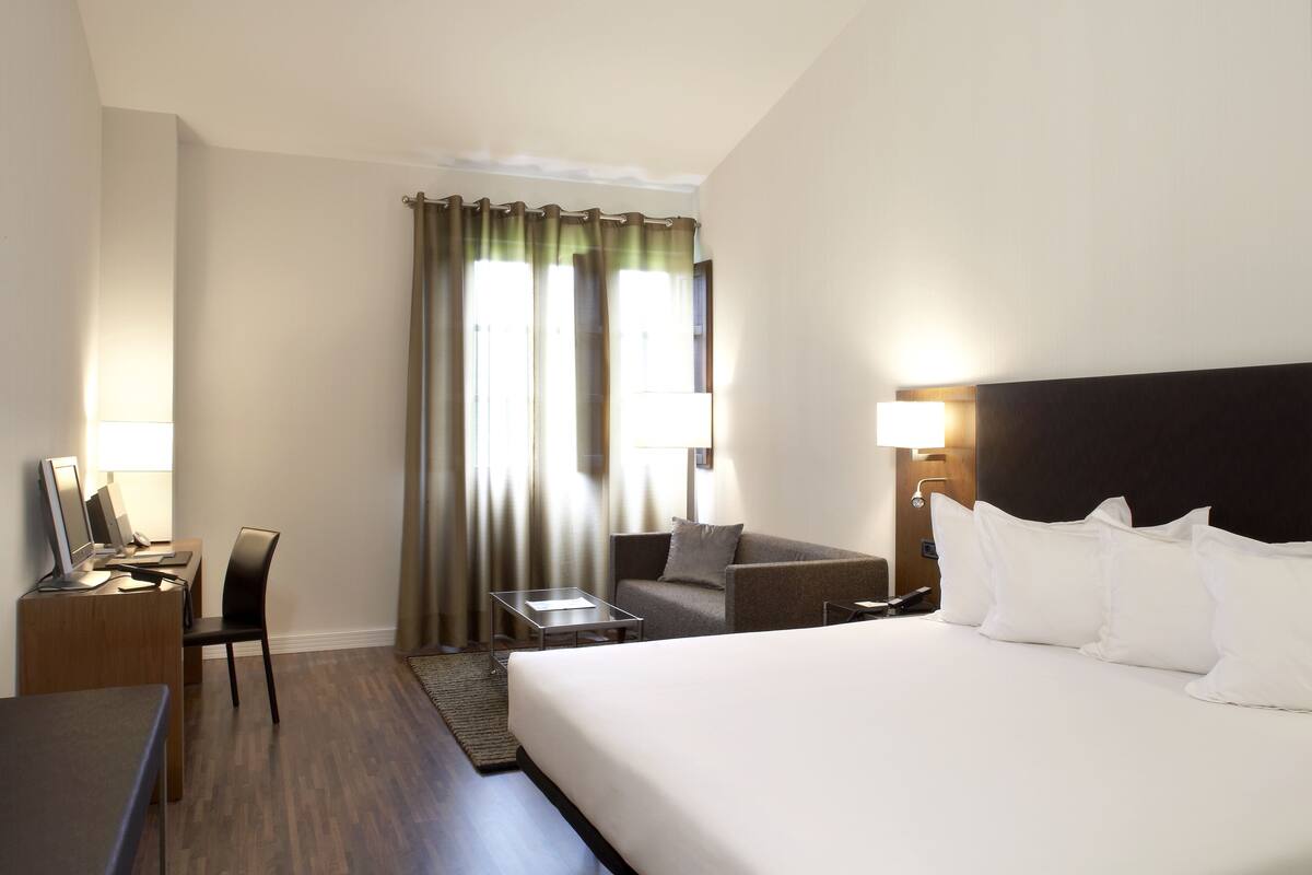 Une chambre d’hôtel avec un lit, un bureau avec un ordinateur, une chaise, un canapé et une fenêtre avec des rideaux.
