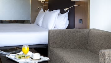 Habitación de hotel con una cama, un sofá gris y una bandeja de desayuno que incluye un vaso de zumo de naranja, una taza y un plato de fruta.