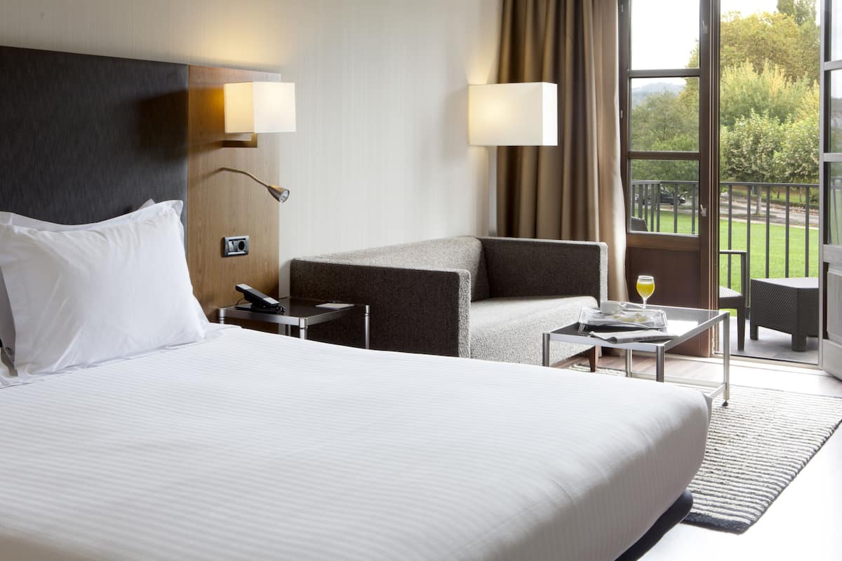 Une chambre d’hôtel avec un grand lit, un canapé, une petite table avec un verre de jus d’orange et un balcon donnant sur des arbres et une voiture.