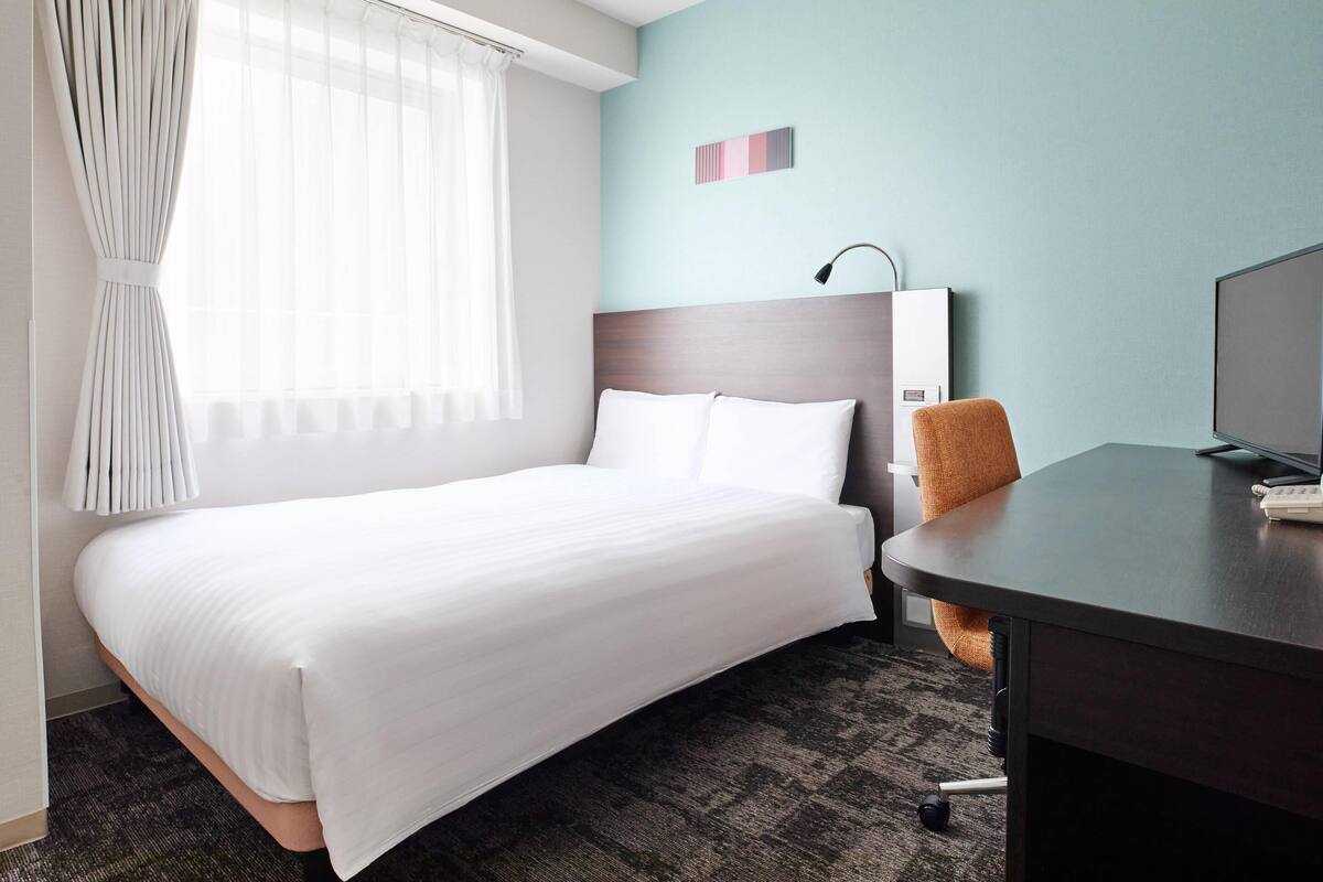 A hotel room with a bed, a desk, a chair, a lamp, and a television.