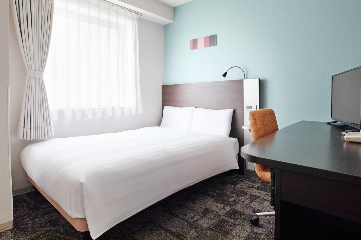A hotel room with a bed, a desk, a chair, a lamp, and a television.