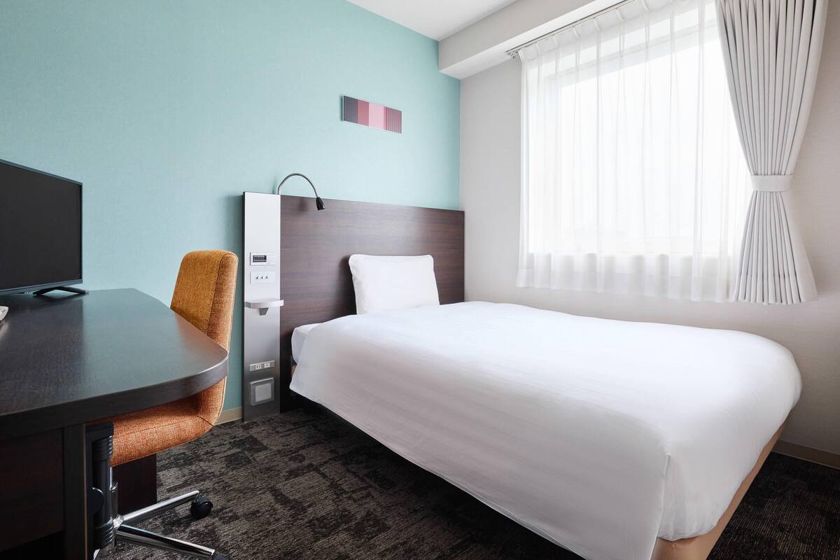 A hotel room with a bed, a desk, a chair, a TV, a wardrobe, and a window with curtains.