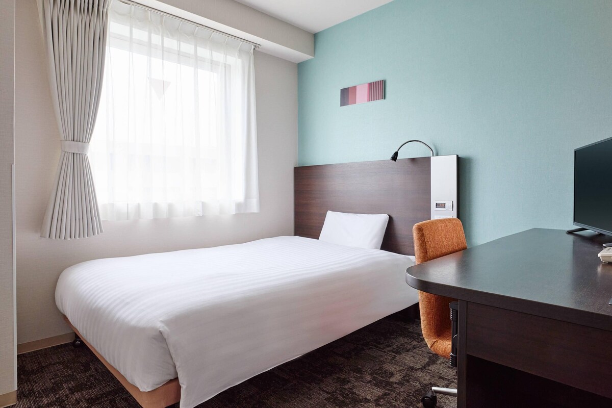 A hotel room with a bed, a desk, a chair, a television, and a window with curtains.