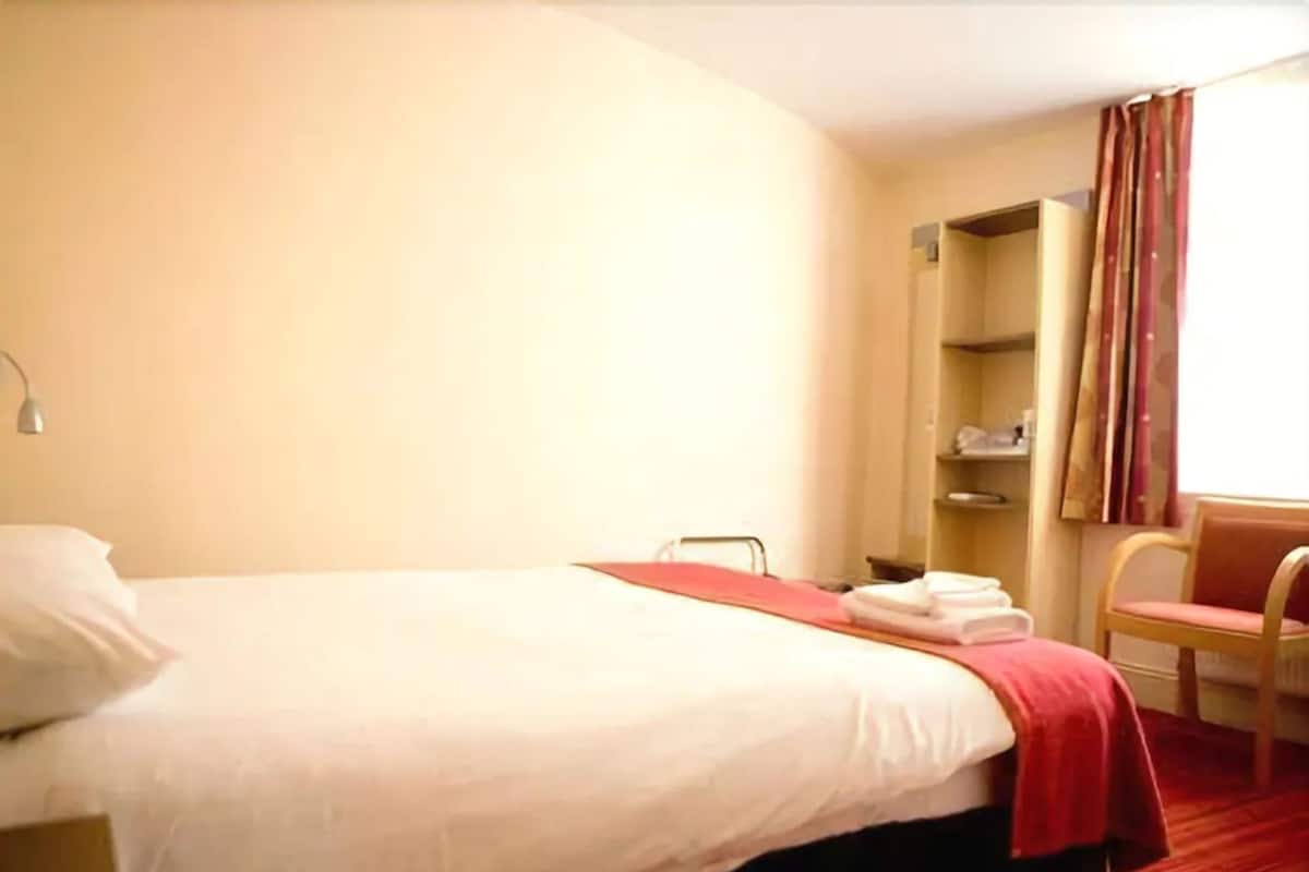 A hotel room with a single bed, a chair, a wardrobe, and a window with curtains.