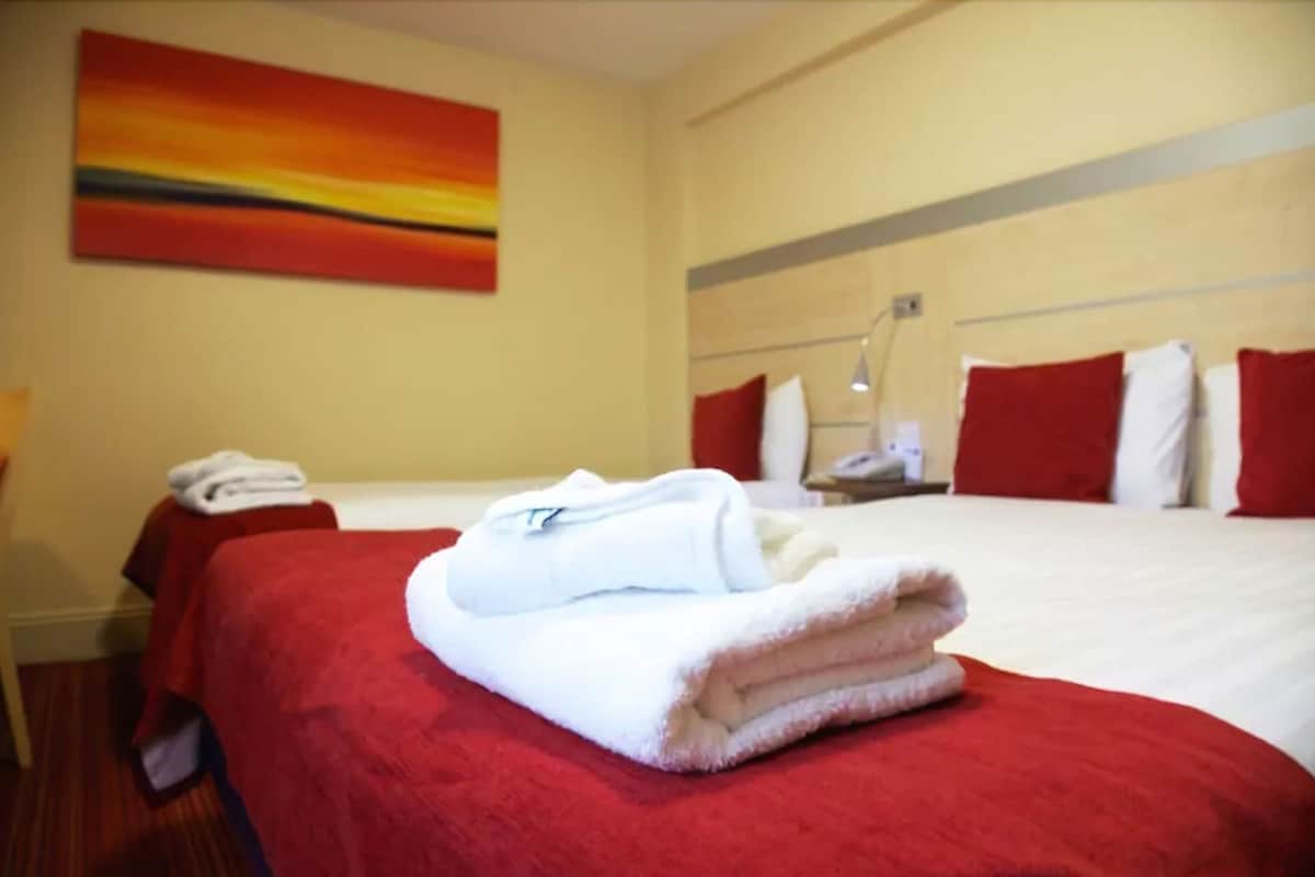 A hotel room with a bed, a red blanket, a white towel, and a painting on the wall.