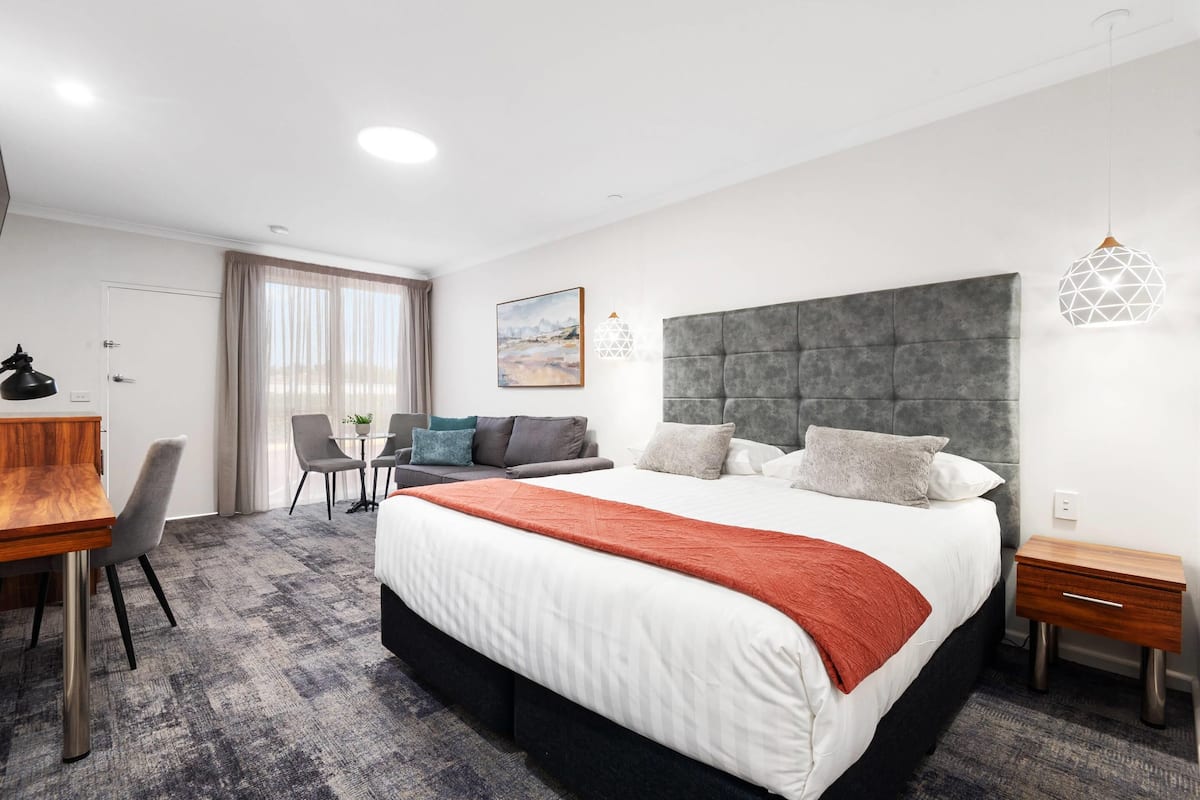 A modern hotel room with a large bed, a desk, a chair, and a dining area.