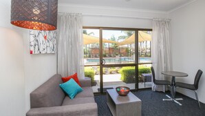 A modern hotel room with a sofa, a coffee table, a chair, and a view of an outdoor swimming pool.
