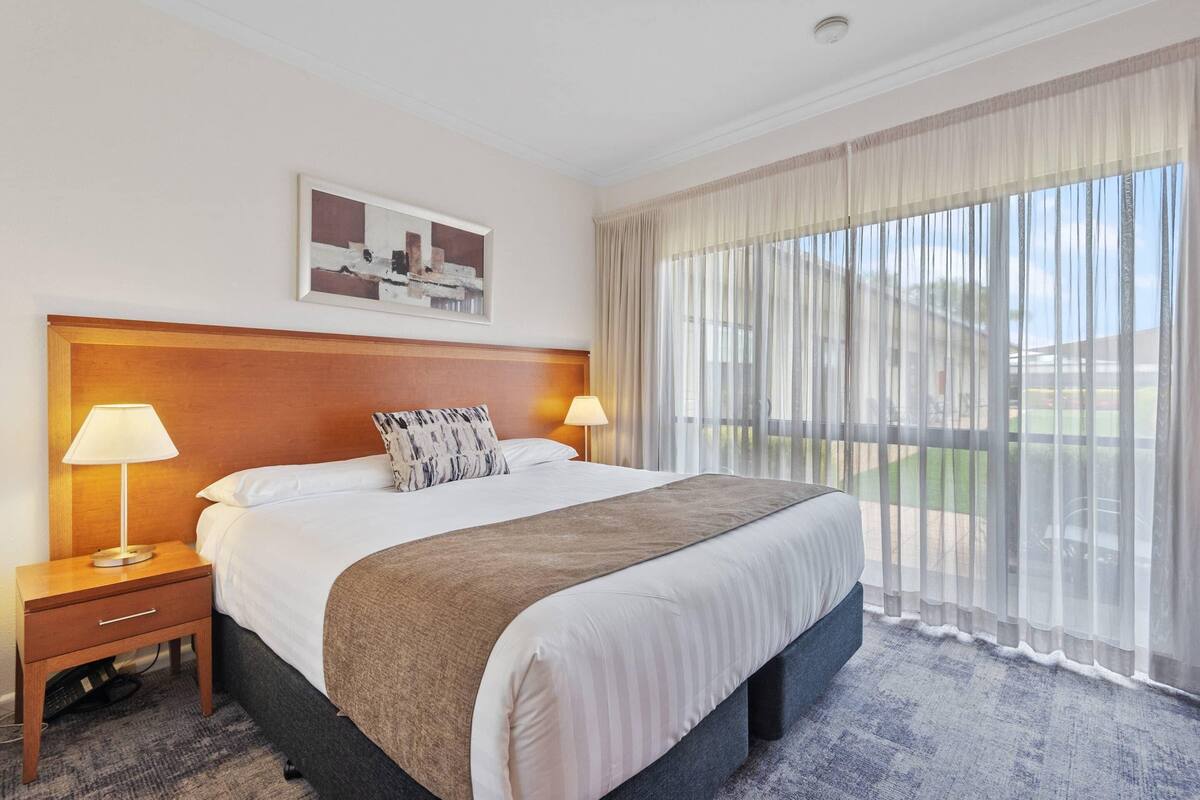 A hotel room with a large bed, two bedside lamps, a headboard, a painting on the wall, and a view of the outdoors through sheer curtains.