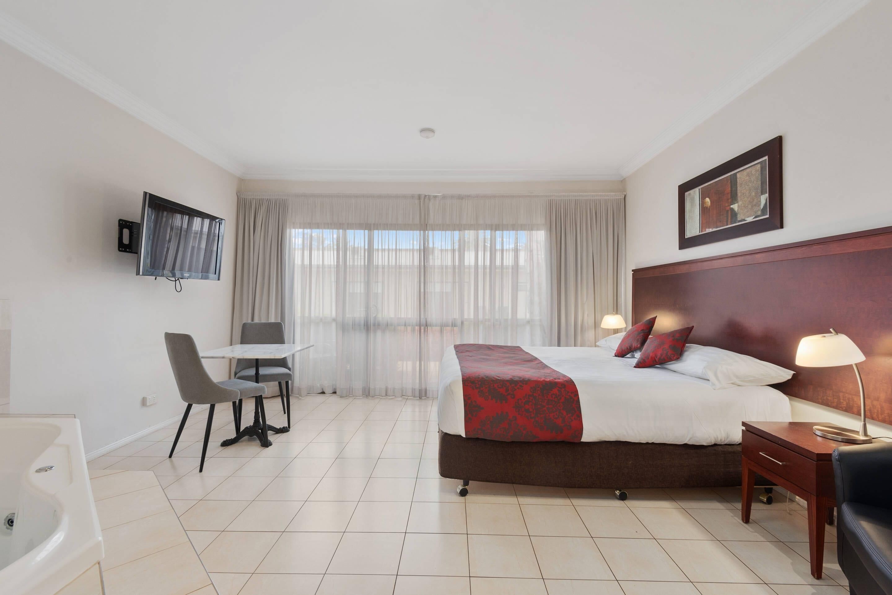 Una habitación de hotel con cama, mesita de noche, lámpara, televisor, silla y una pequeña mesa.