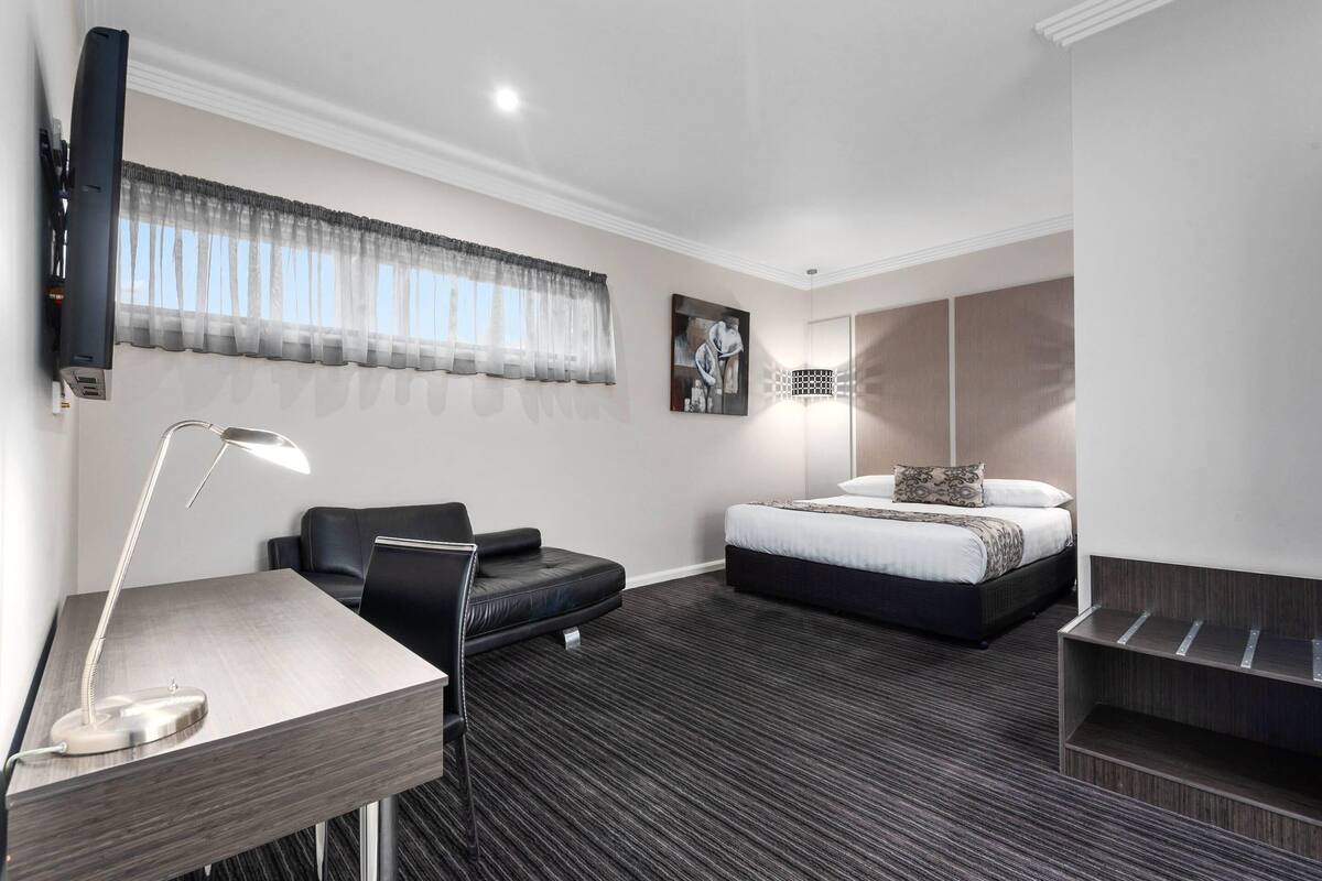 A modern hotel room with a bed, a desk with a lamp, a chair, a sofa, and a large window with sheer curtains.
