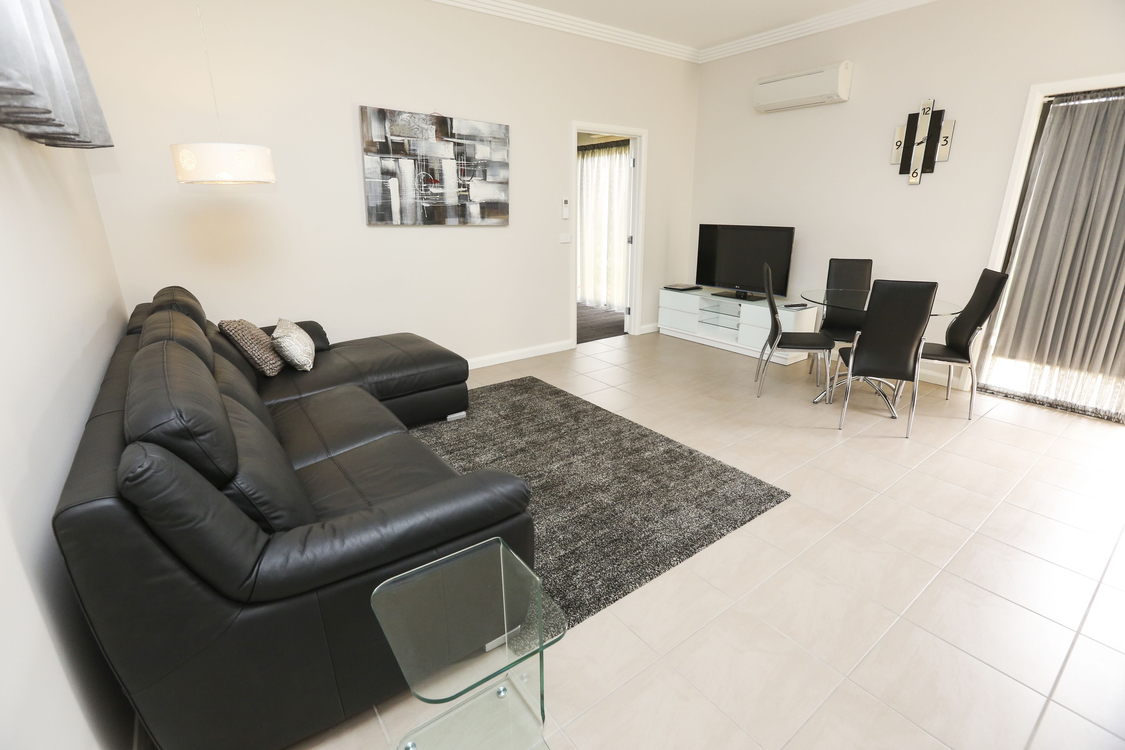 Un salon moderne avec un canapé en cuir noir, une table basse en verre, un téléviseur à écran plat et un coin repas comprenant des chaises et un buffet blanc.