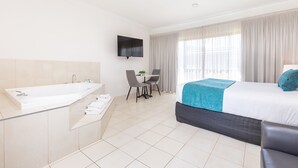 A modern hotel room with a large bathtub, a dining area with a table and chairs, a bed with a blue blanket, and a TV mounted on the wall.