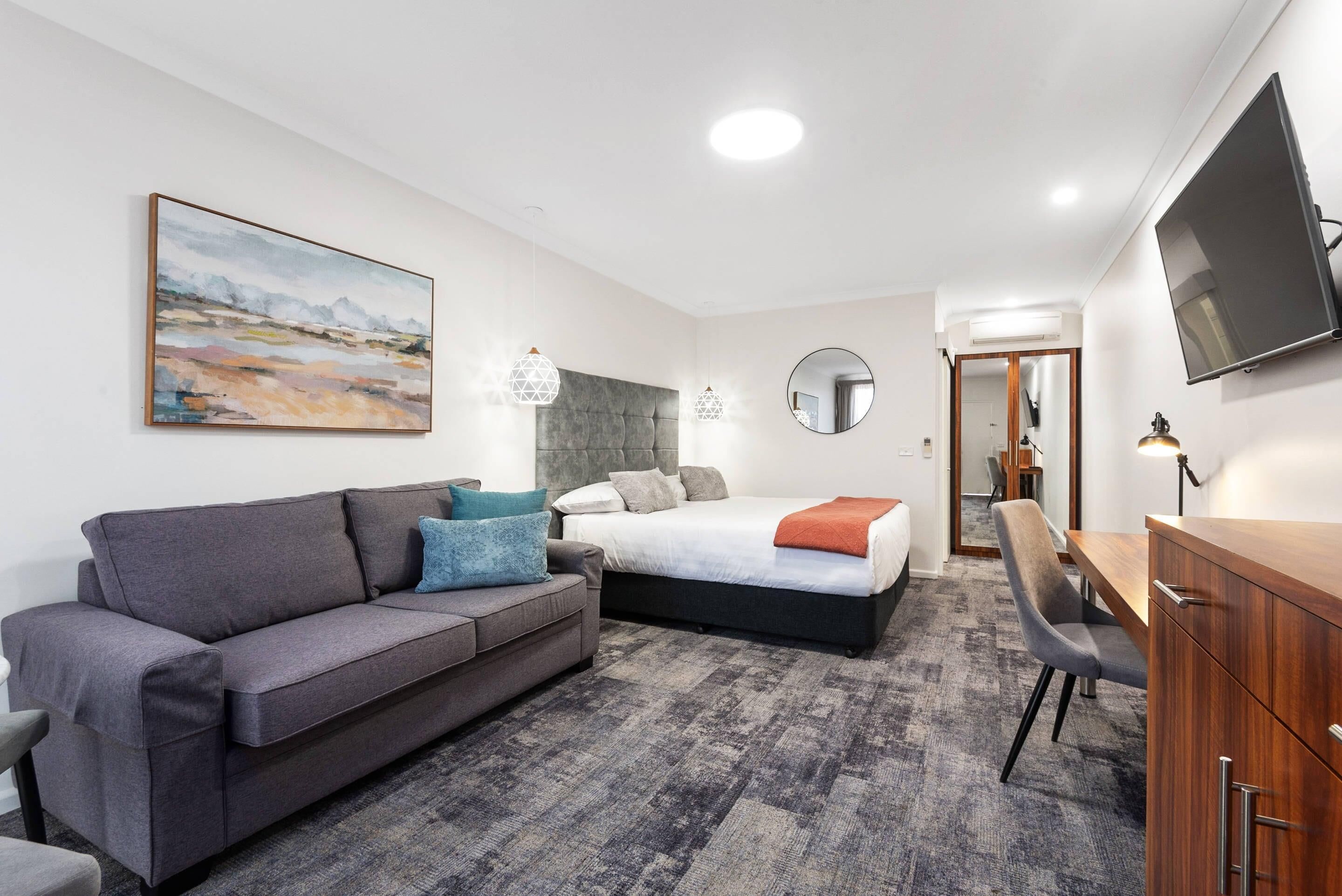 Una habitación de hotel moderna con un sofá gris, una cama con cabecera gris, un escritorio y un televisor montado en la pared.