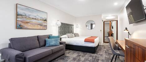 Una habitación de hotel moderna con un sofá gris, una cama con cabecera gris, un escritorio y un televisor montado en la pared.