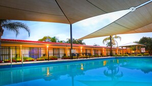 A swimming pool with a covered area, palm trees, and a building with a red roof in the background.