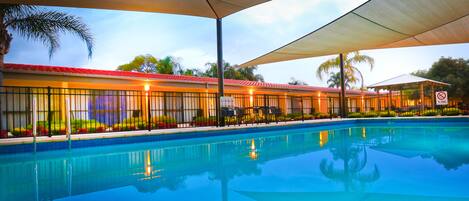 Una piscina con zona techada, palmeras y un edificio de techo rojo al fondo.