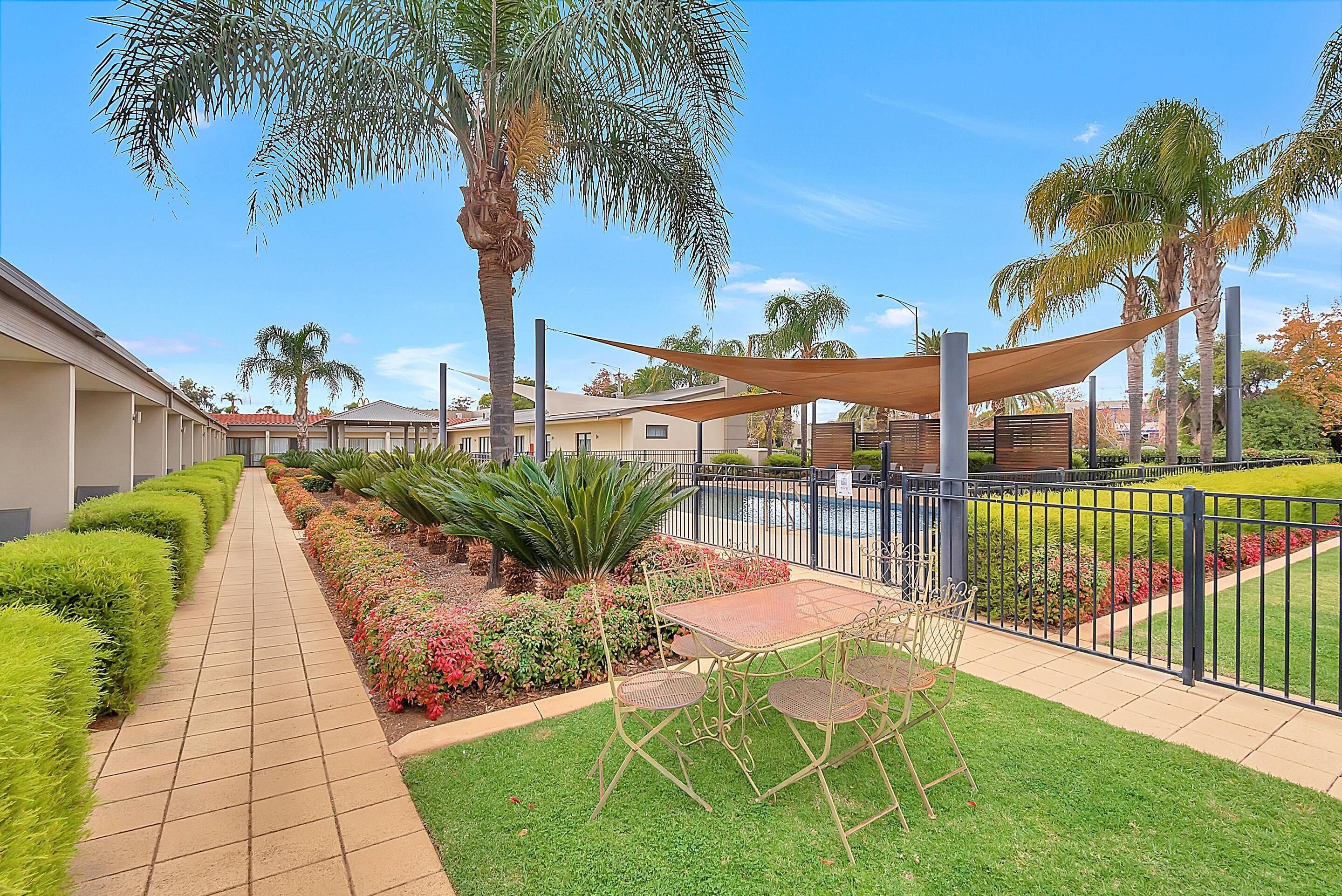 A courtyard with a paved walkway, a shaded table and chairs, and a variety of plants and flowers.
