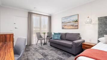 Habitación de hotel con un sofá gris, dos sillones grises, un escritorio de madera, una mesita con una planta, un cuadro en la pared y una lámpara colgante de diseño geométrico.