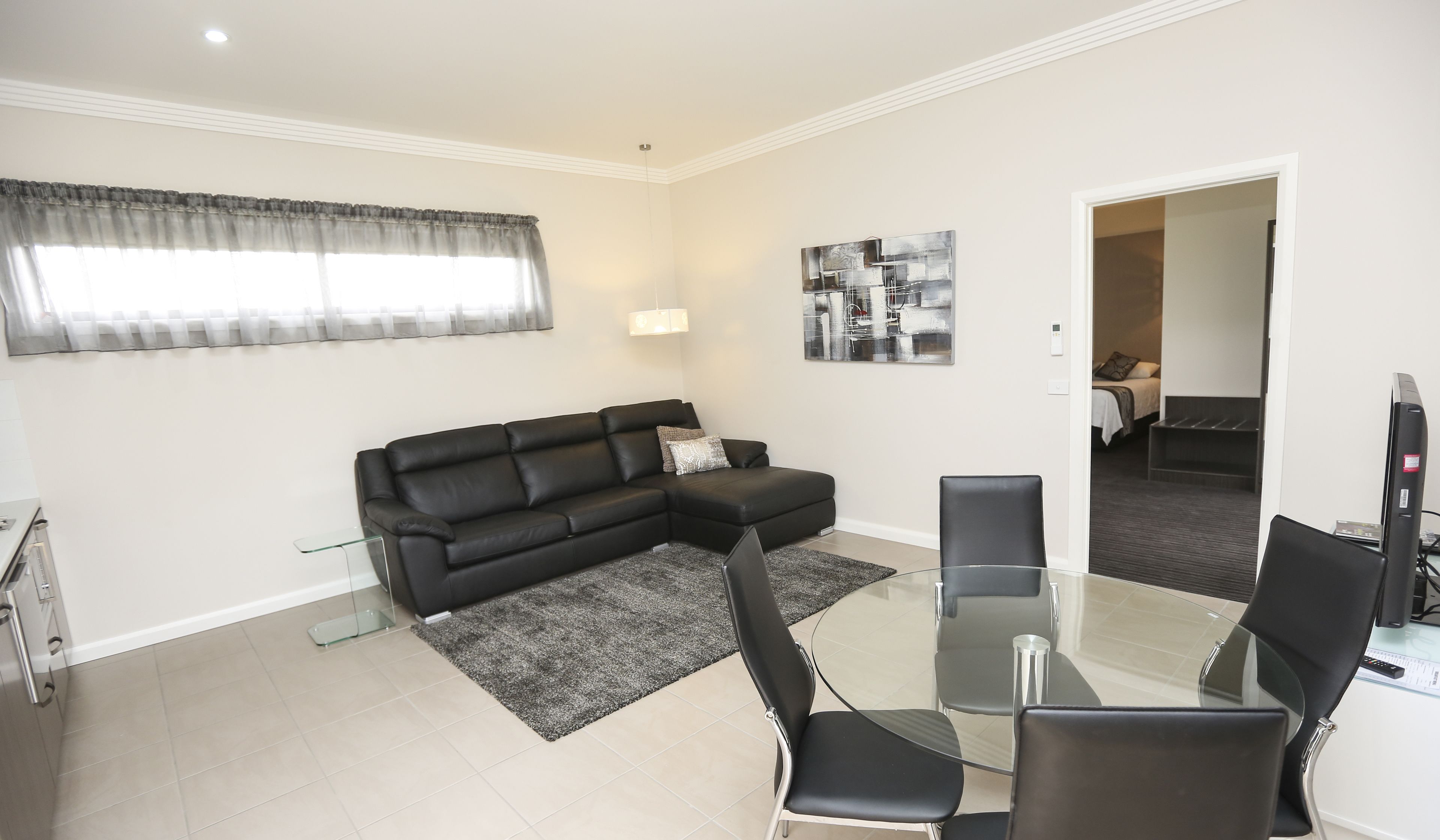 A modern living room with a black leather sofa, a glass coffee table, and black chairs. There is a window with sheer curtains, a painting on the wall, and a door leading to another room.