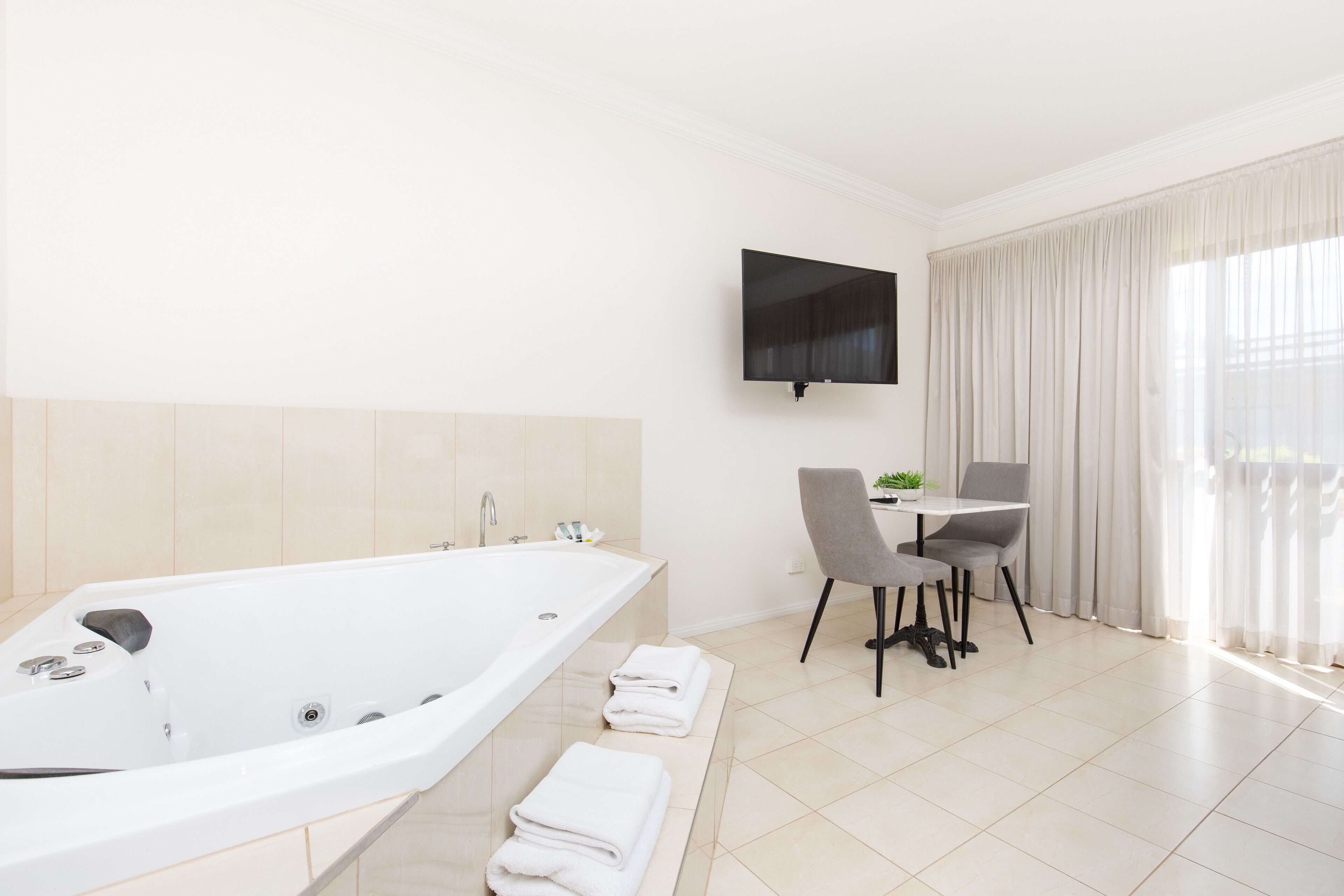 A modern bathroom with a large bathtub, a flat-screen TV mounted on the wall, and a dining area with two chairs and a small table.