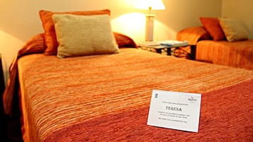Hotellihuoneessa on kaksi sänkyä, joissa on oranssit vuodevaatteet ja tyynyt. Valkoinen kyltti, jossa lukee nimi Teresa, on asetettu toisen sängyn päälle.