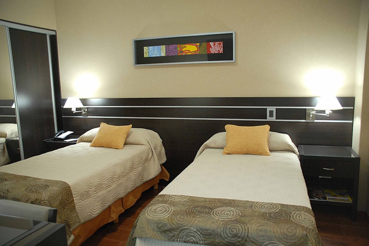 A hotel room with two beds, each with a pillow and a bedside table with a lamp. There is a framed artwork on the wall above the beds.
