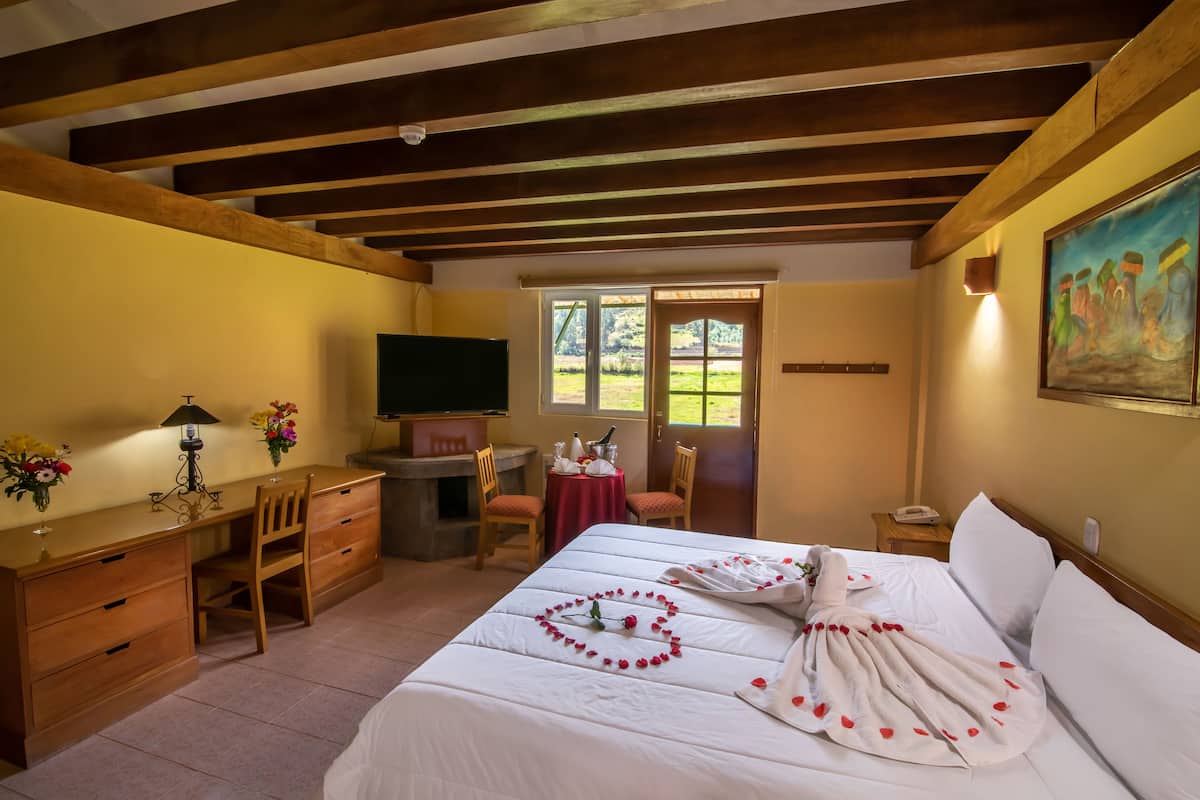 Une chambre d’hôtel avec un plafond en bois, un lit parsemé de pétales de rose, un bureau avec une lampe, une télévision et un tableau accroché au mur.