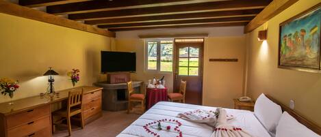 A hotel room with a wooden ceiling, a bed with rose petals, a desk with a lamp, a TV, and a painting on the wall.