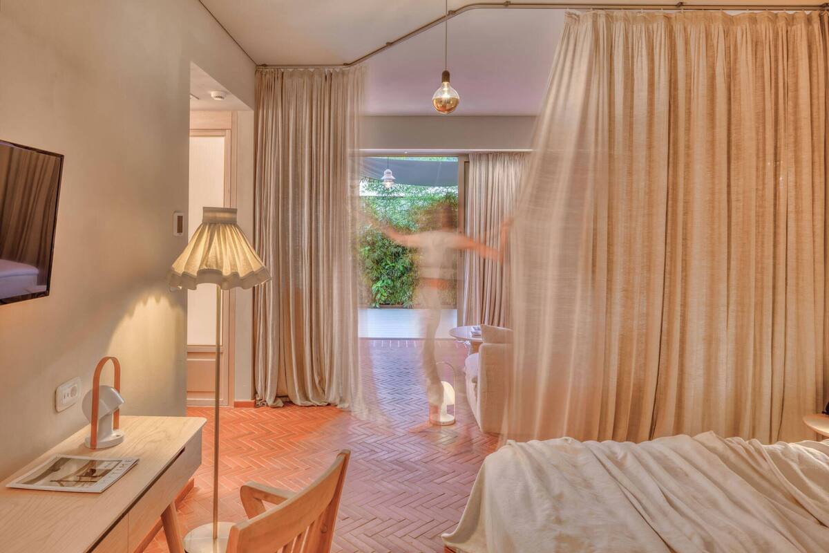 A hotel room with a bed, a desk, a lamp, and a view of greenery outside.