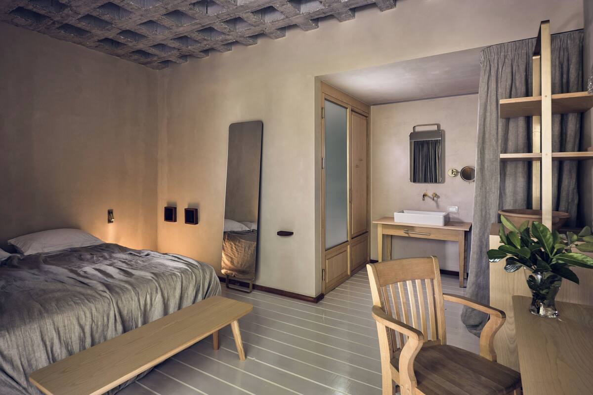 A bedroom with a bed, a bedside table, a chair, a mirror, a wardrobe, and a small bathroom area with a sink and a mirror.