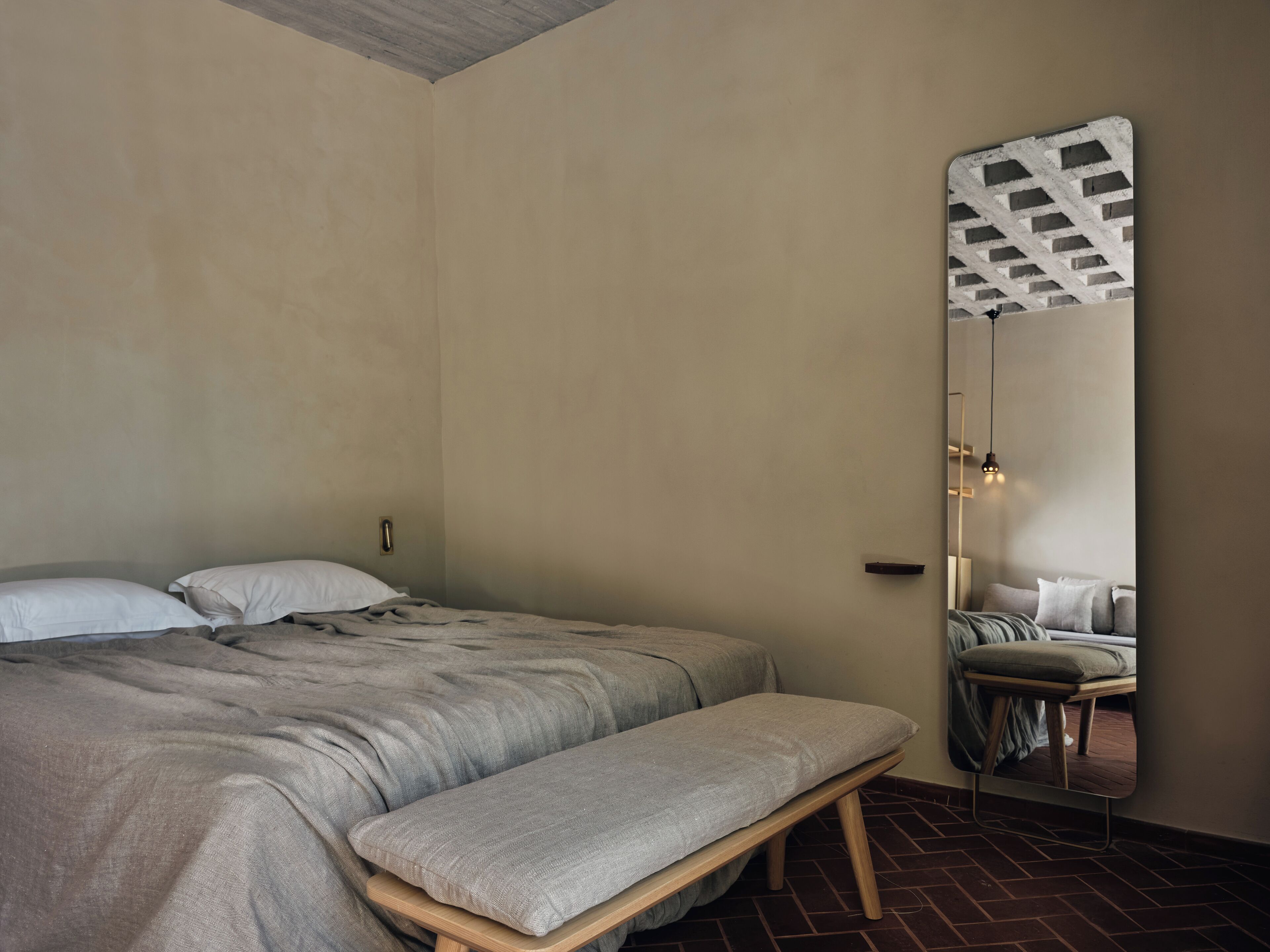 A bedroom with a bed, a bench, a mirror, and a brick floor.