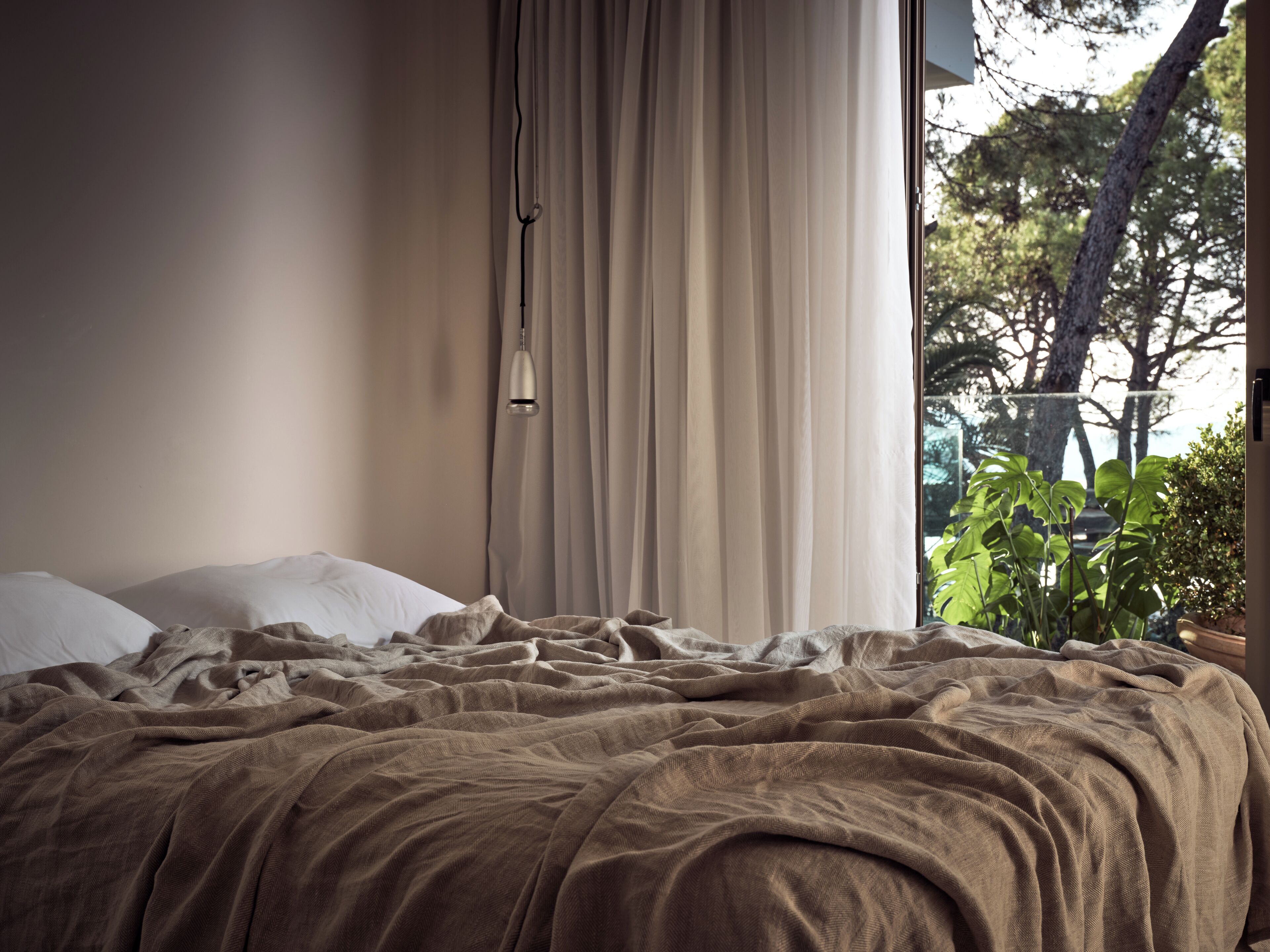 A bedroom with a bed, curtains, a hanging light, and a view of trees through a window.