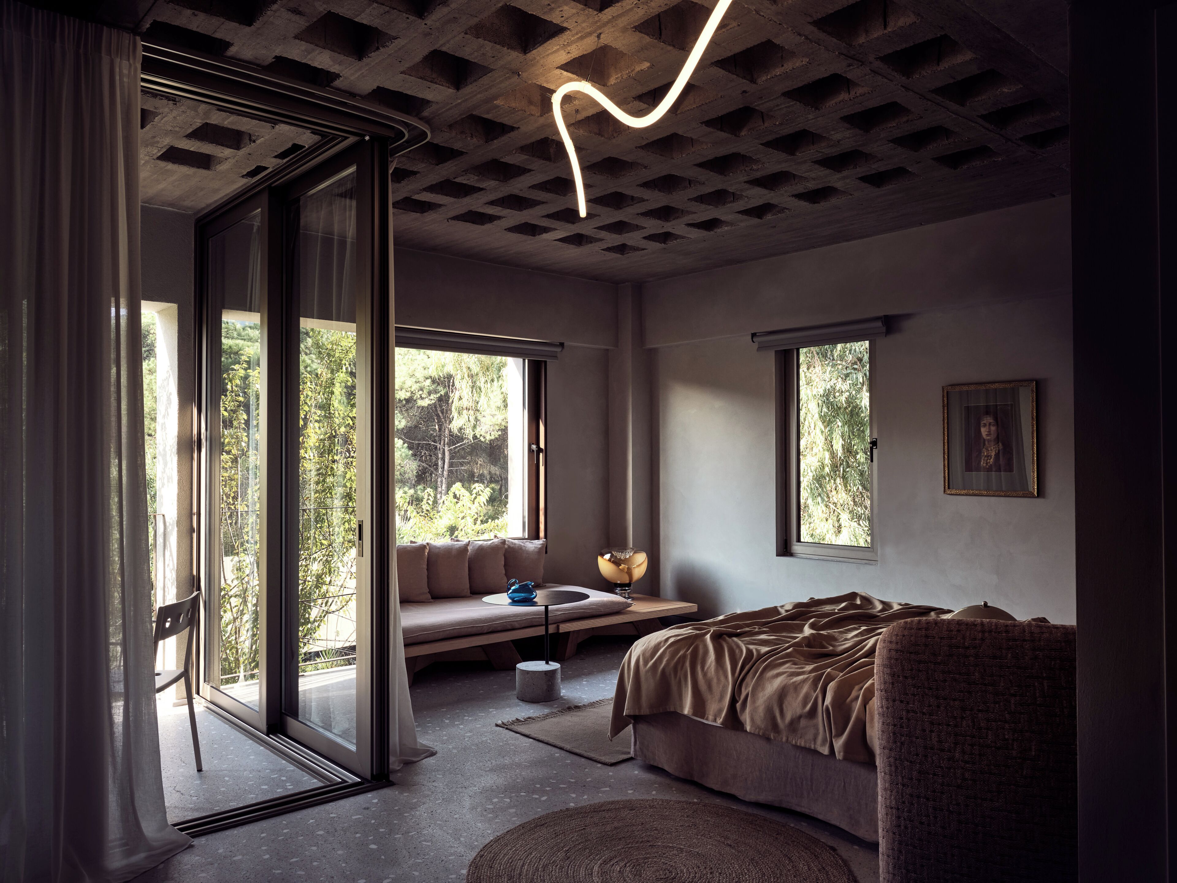 A modern bedroom with a large bed, a sofa, and a desk. There are two windows providing natural light and a view of greenery outside.