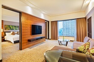 Una habitación de hotel moderna con una cama grande, un televisor de pantalla plana montado en un panel de madera, una mesa de centro de vidrio, un sofá y un ventanal con cortinas.