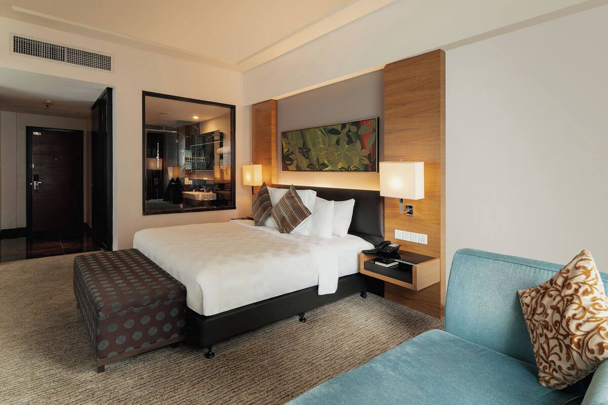 Een hotelkamer met een bed, nachtkastje, lamp, bank en grote spiegel.