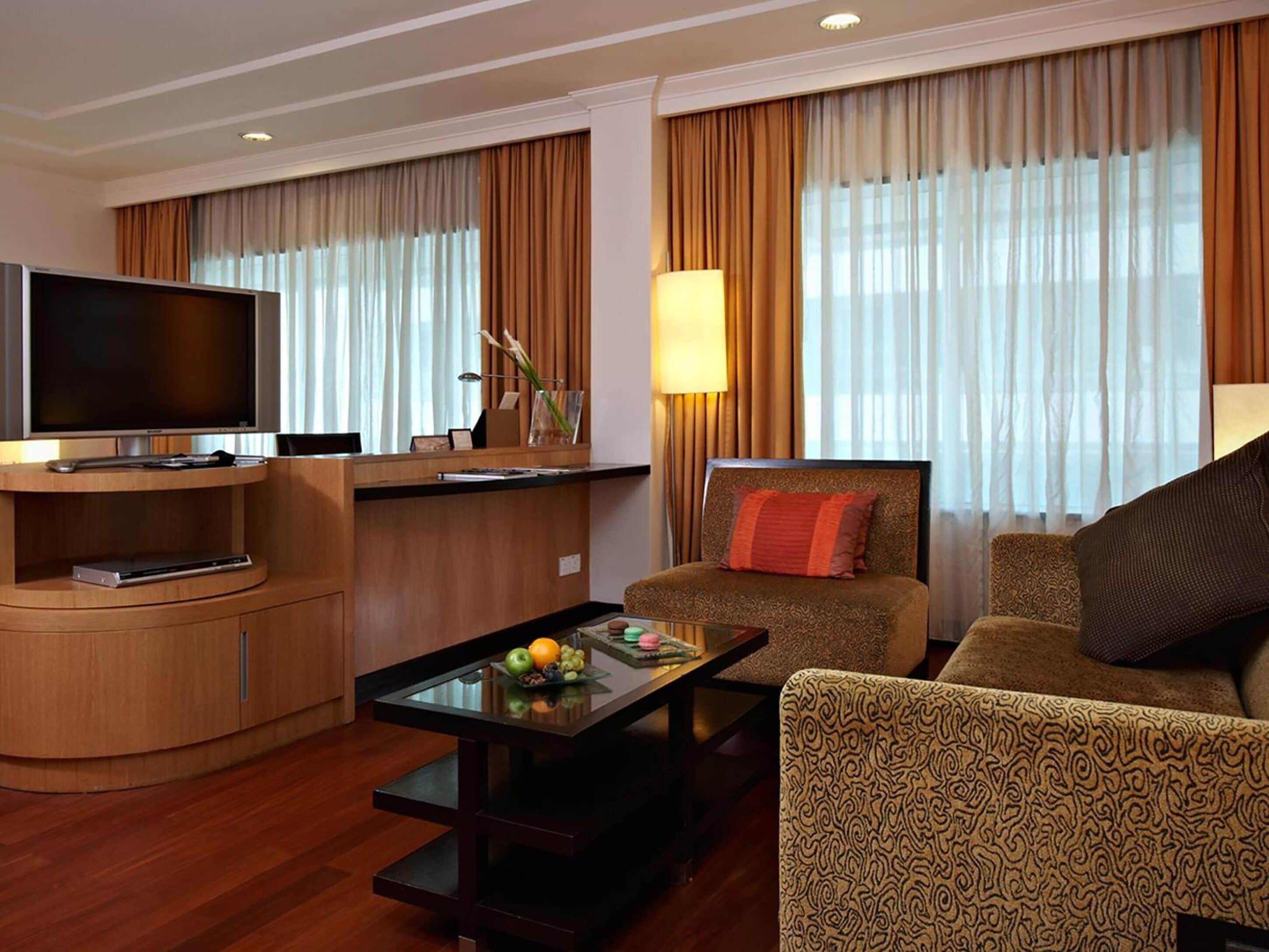 Habitación de hotel moderna con televisor de pantalla plana, una mesa de centro de vidrio con fruta, un sofá y una silla.