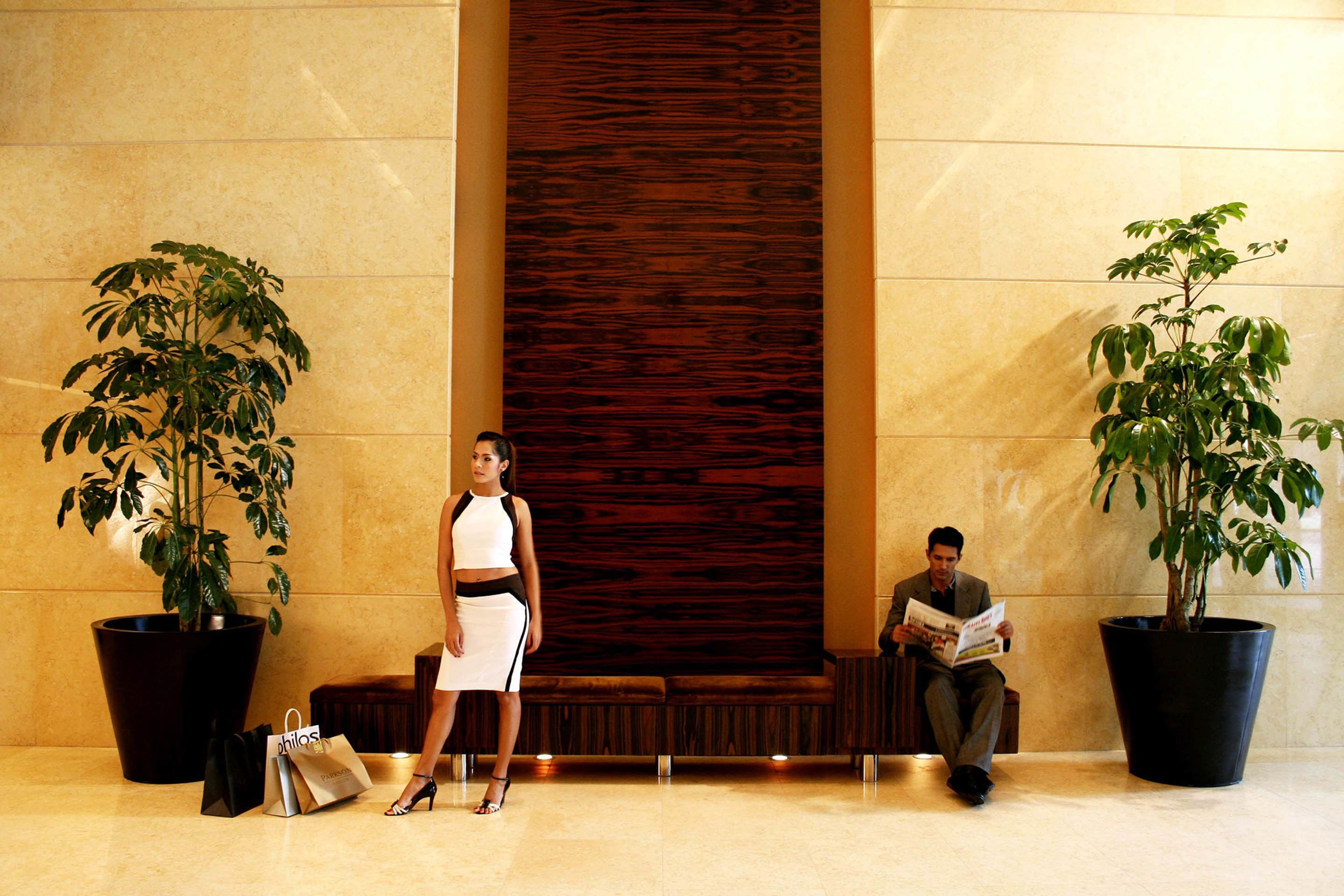 A lobby with a large wooden panel, two potted plants, a bench, and a woman standing.