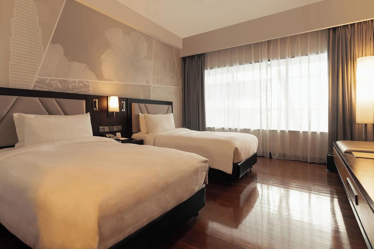 Een hotelkamer met twee bedden, een nachtkastje met een lamp, een groot raam met gordijnen en een stadsmuur op de muur.