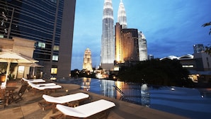 Zona de piscina en la azotea con sillones de descanso y sombrillas, que refleja edificios cercanos, incluyendo las Torres Petronas.