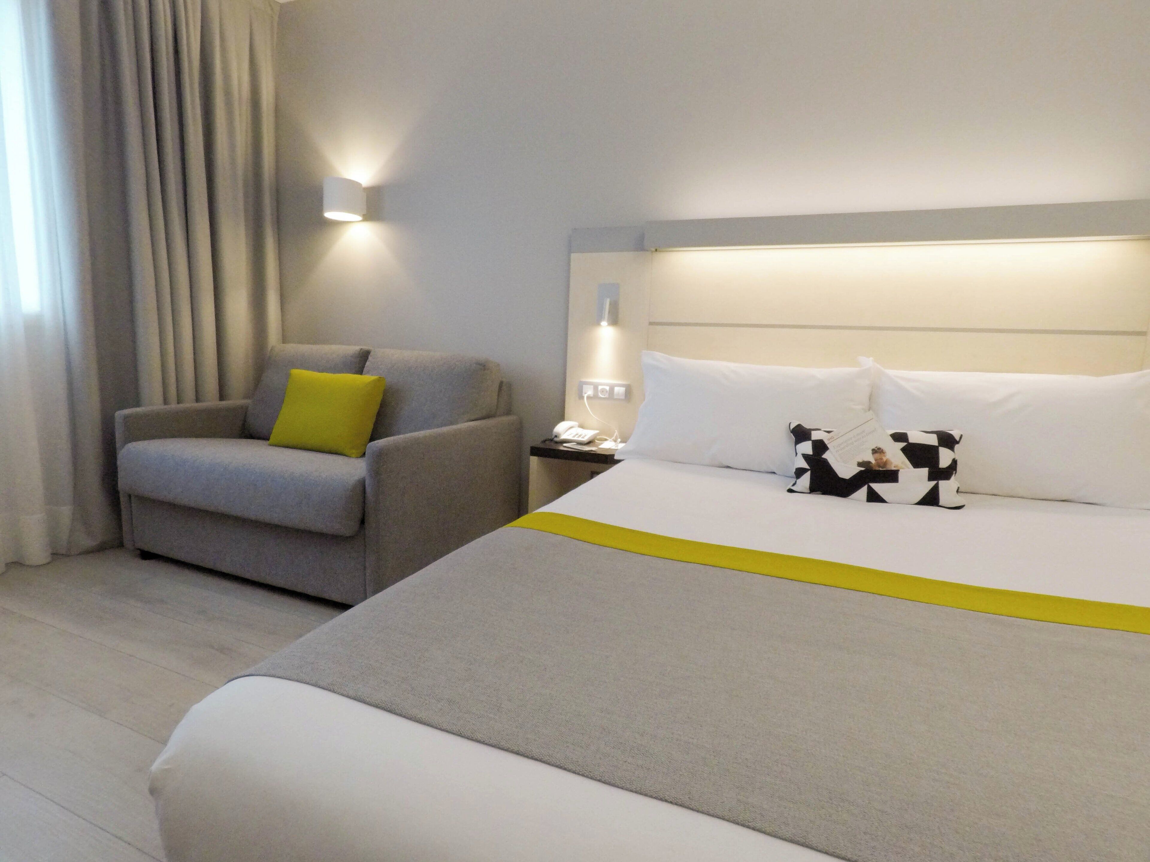 A hotel room with a bed, a chair, a nightstand, and a wall-mounted lamp.
