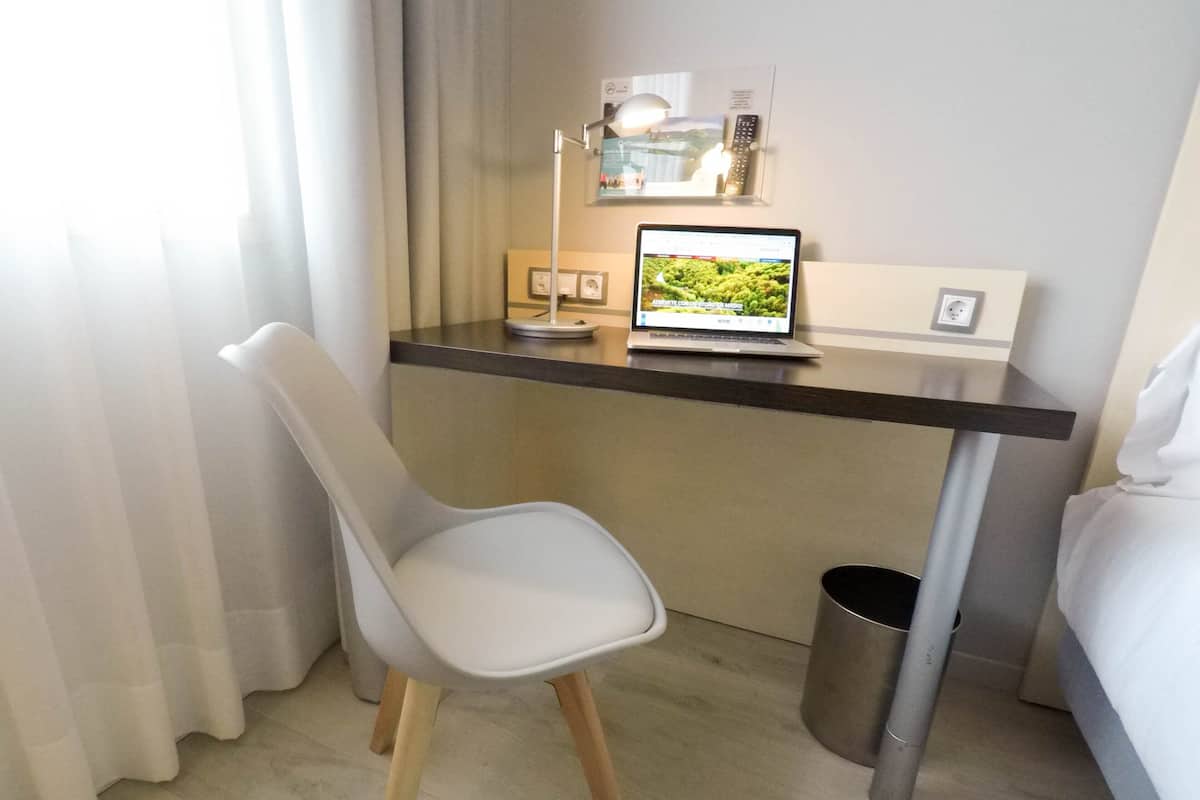 A small, modern hotel room with a desk, a laptop, a chair, a bed, and a trash bin.